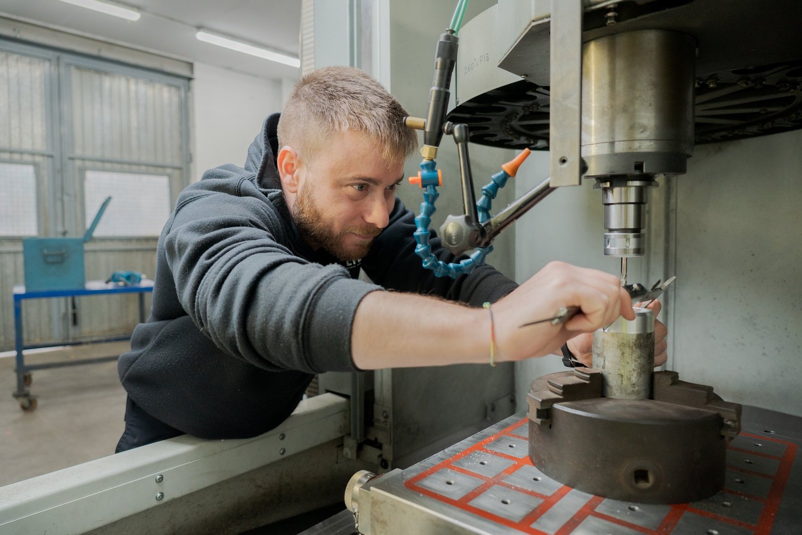 Hombre barbudo en sudadera negra ajusta una pieza metálica en una máquina industrial de mecanizado.