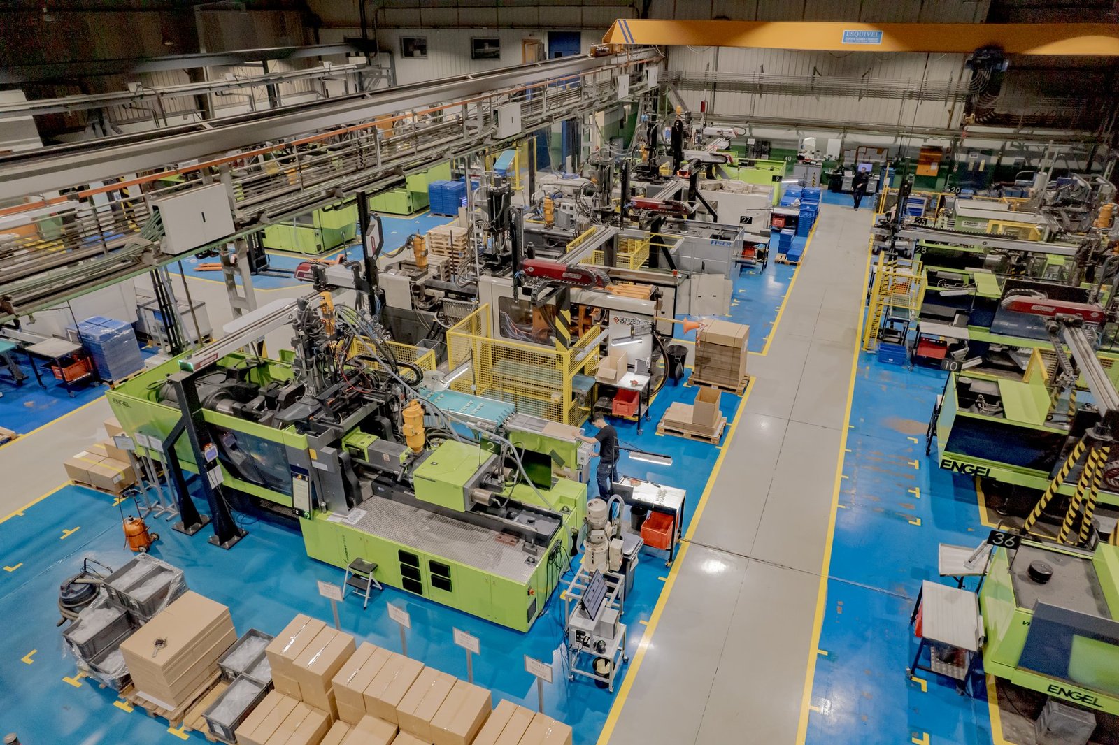 Vista aérea de fábrica industrial MOLTEC. Múltiples máquinas de inyección, robots, operarios y palets con cajas. Suelo azul y gris.