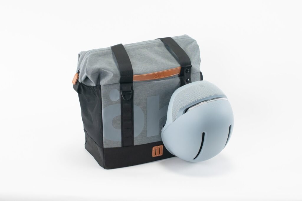 Bolso de tela gris y negro con asas de cuero marrón y logo Öhll, junto a un casco azul claro con rejilla superior.