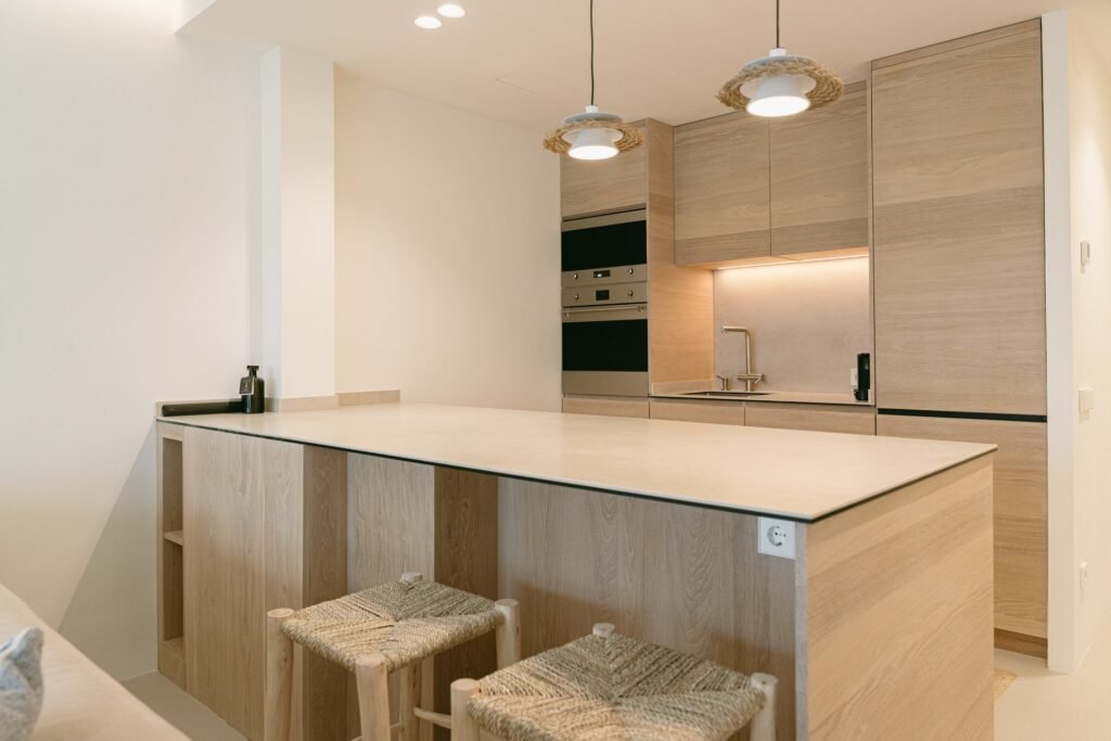 Cocina moderna con isla de madera clara, encimera beige, dos taburetes de esparto y lámparas colgantes.