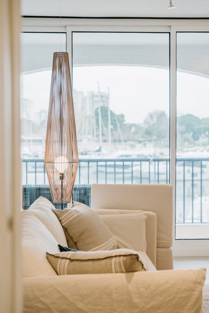 Lámpara colgante de diseño moderno con pantalla de lamas de madera, iluminada, sobre un sofá claro con cojines. Al fondo, un puerto con b…
