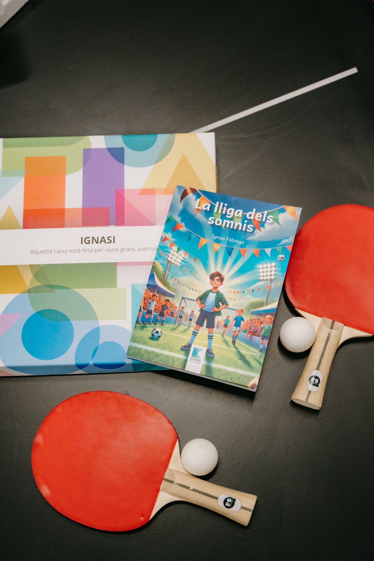 Caja KALEIDOBOOK IGNASI con diseño geométrico, libro 'La lliga dels somnis' y dos palas de ping-pong con bolas.