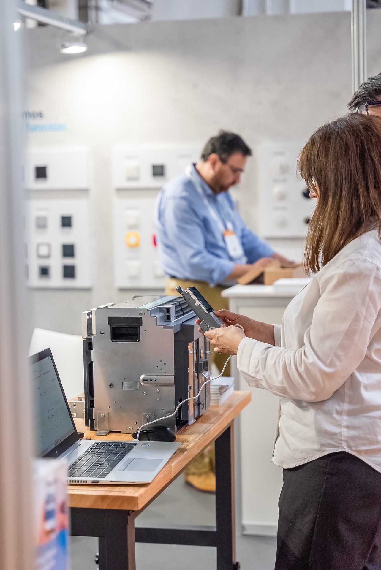 Mujer conecta dispositivo a equipo en stand Efintec, con portátil en mesa. Hombre en segundo plano junto a exposición de productos.