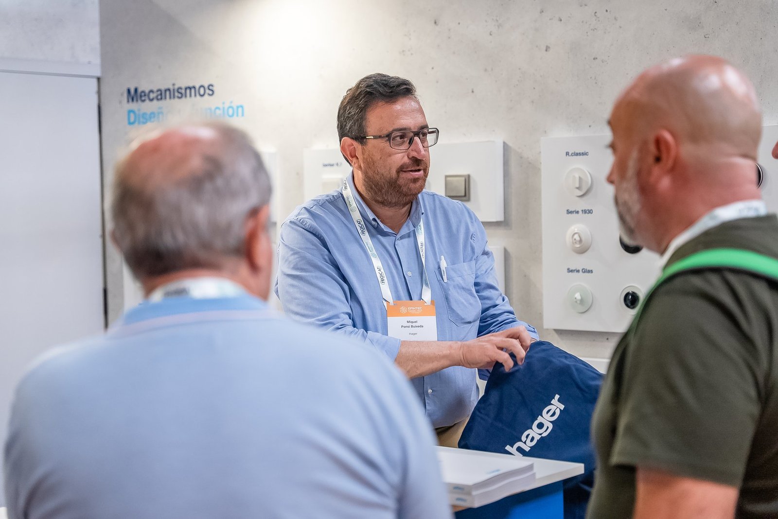 Miquel Ponsí Bulseda de Hager en Efintec, conversando con dos asistentes en el stand. Se aprecian mecanismos eléctricos.