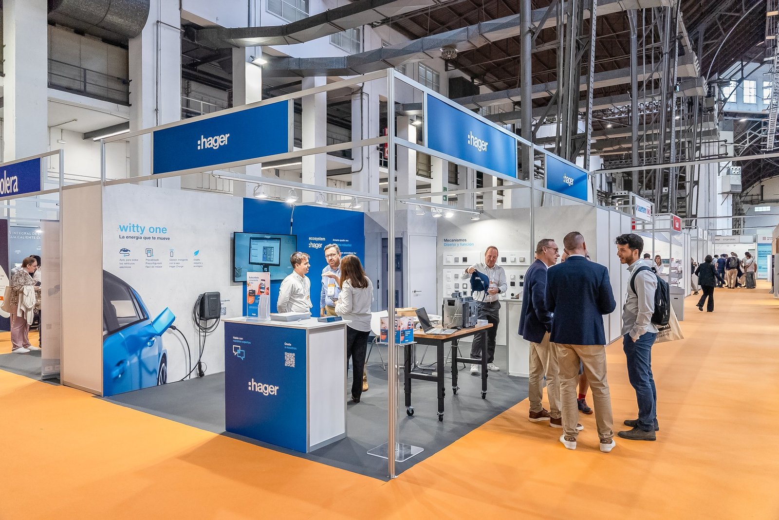 Stand de Hager en Efintec con visitantes interactuando, mostrando cargadores de coche eléctrico y mecanismos eléctricos.