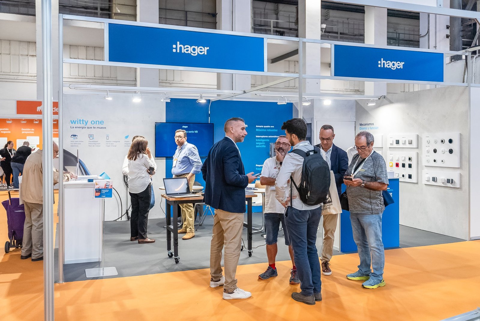 Stand de Hager en Efintec con paneles azules, personas conversando y exhibiendo productos eléctricos como cargadores EV y mecanismos.