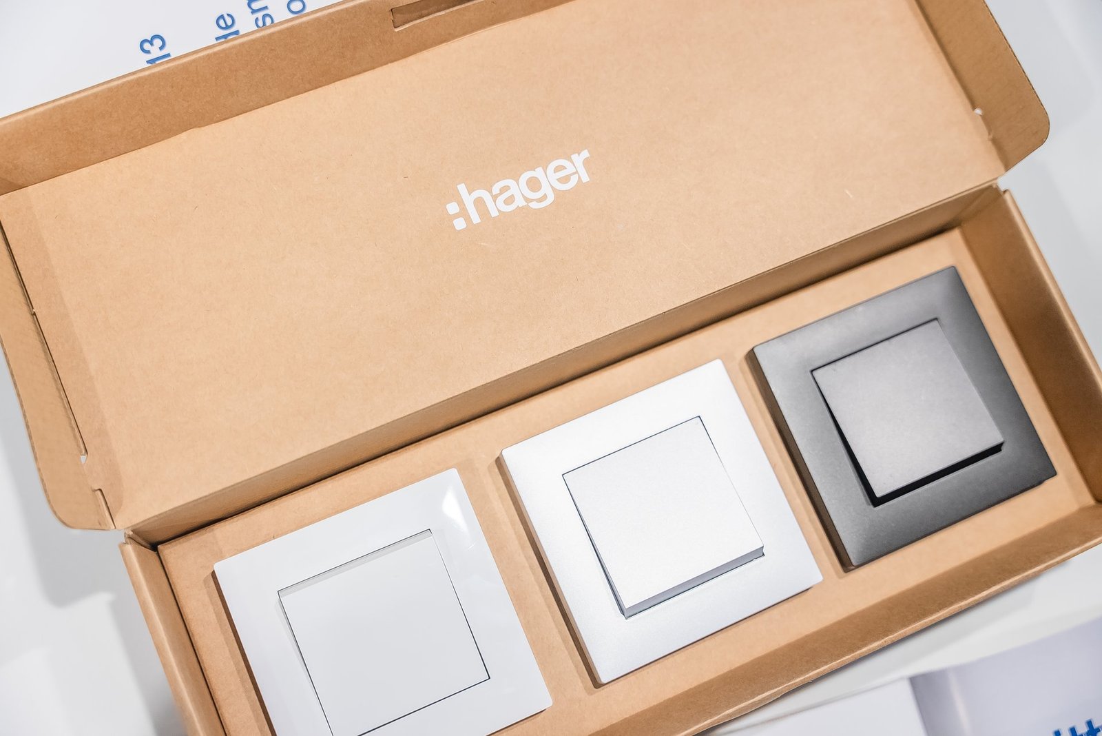 Caja abierta con el logo de Hager, mostrando tres interruptores modernos en blanco, plata y negro. Muestras de producto.