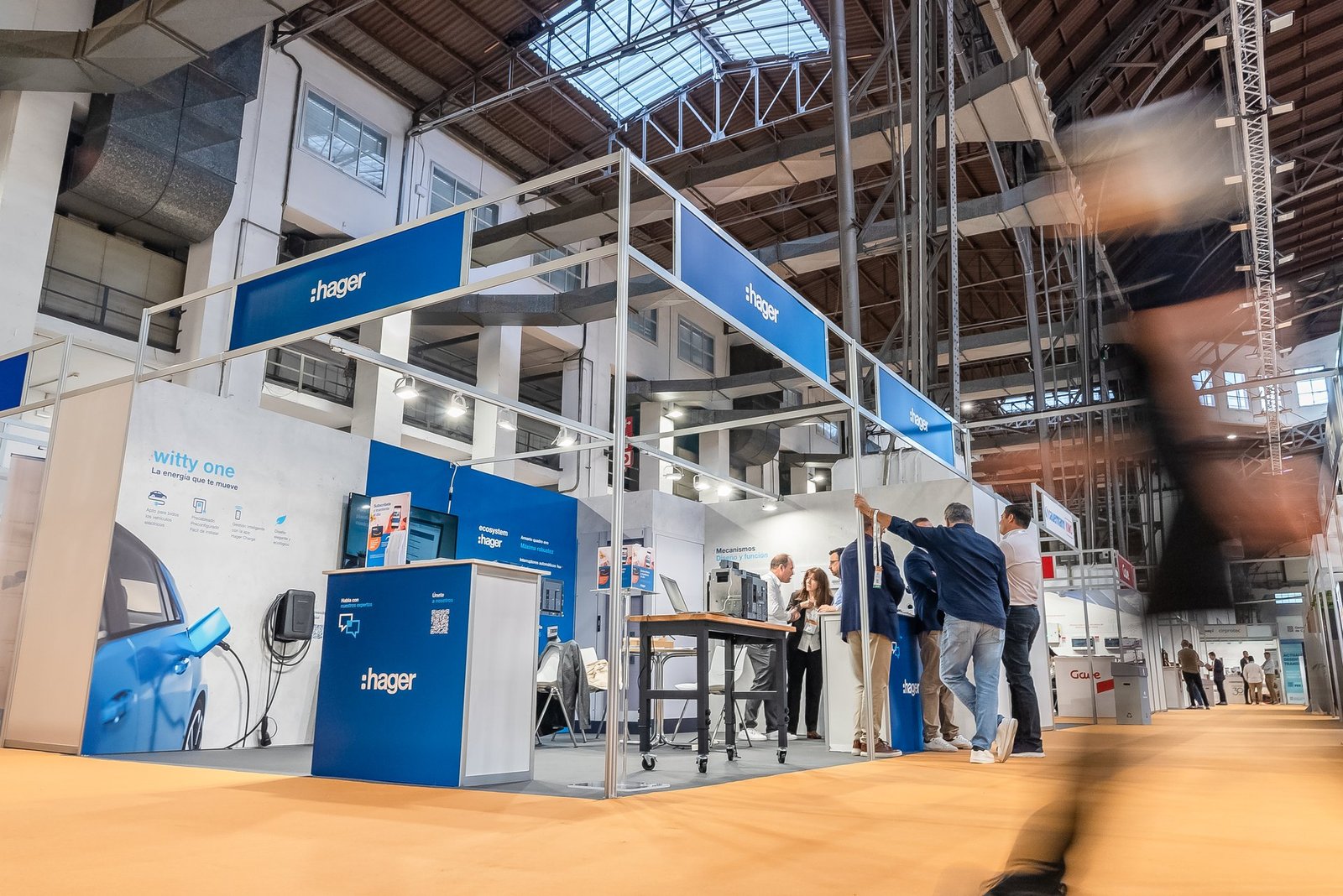 Stand de Hager en Efintec con asistentes. Muestra soluciones de carga de vehículos eléctricos y productos. Techo industrial.