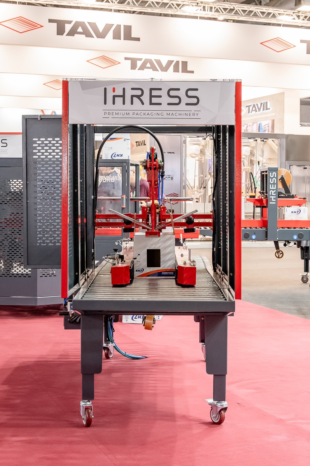 Máquina de embalaje IHRESS roja y gris con una caja en la cinta transportadora, en el stand de Hispack.