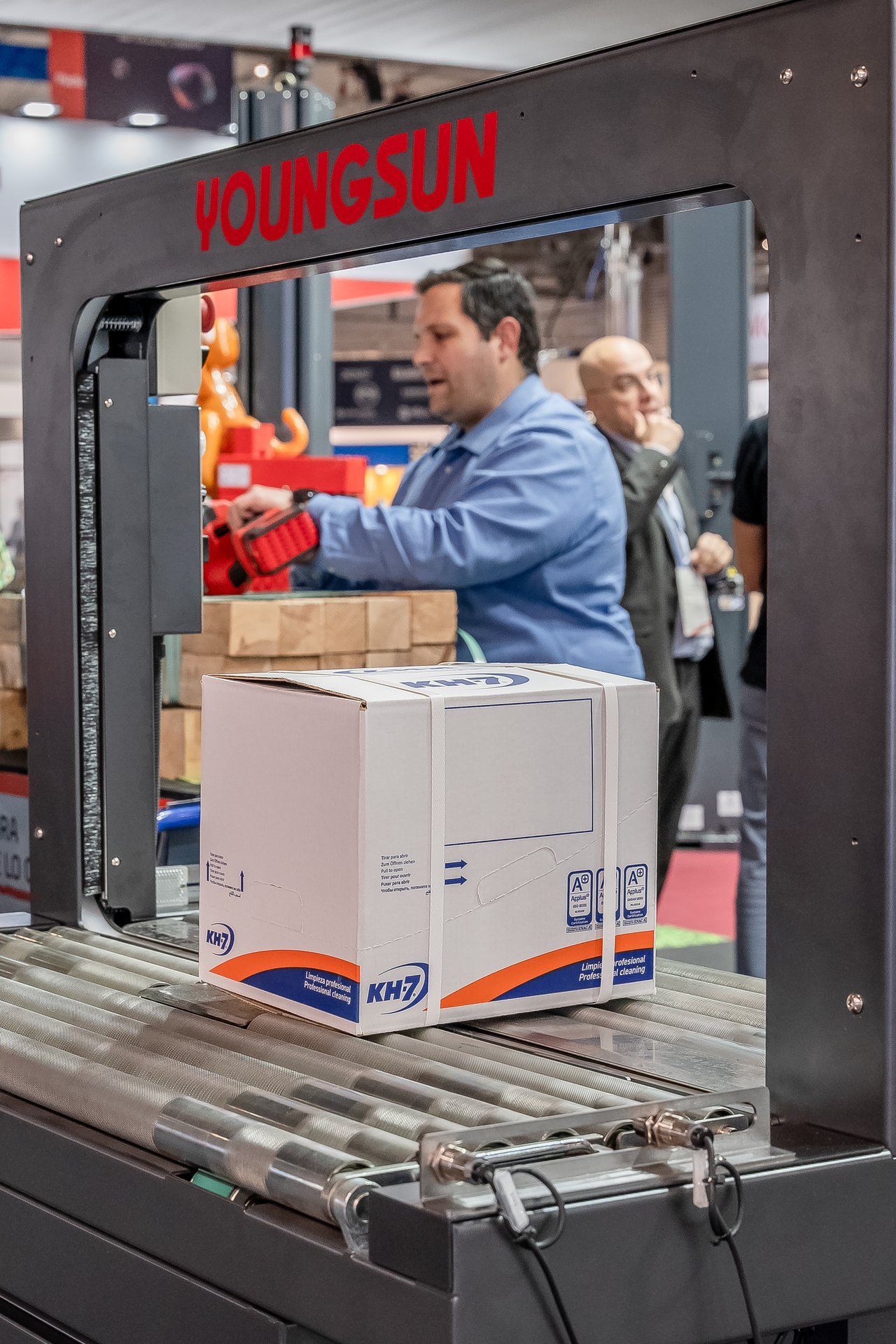 Caja KH7 en máquina flejadora Youngsun en Hispack. Un operario ajusta el fleje, otro observa el proceso.