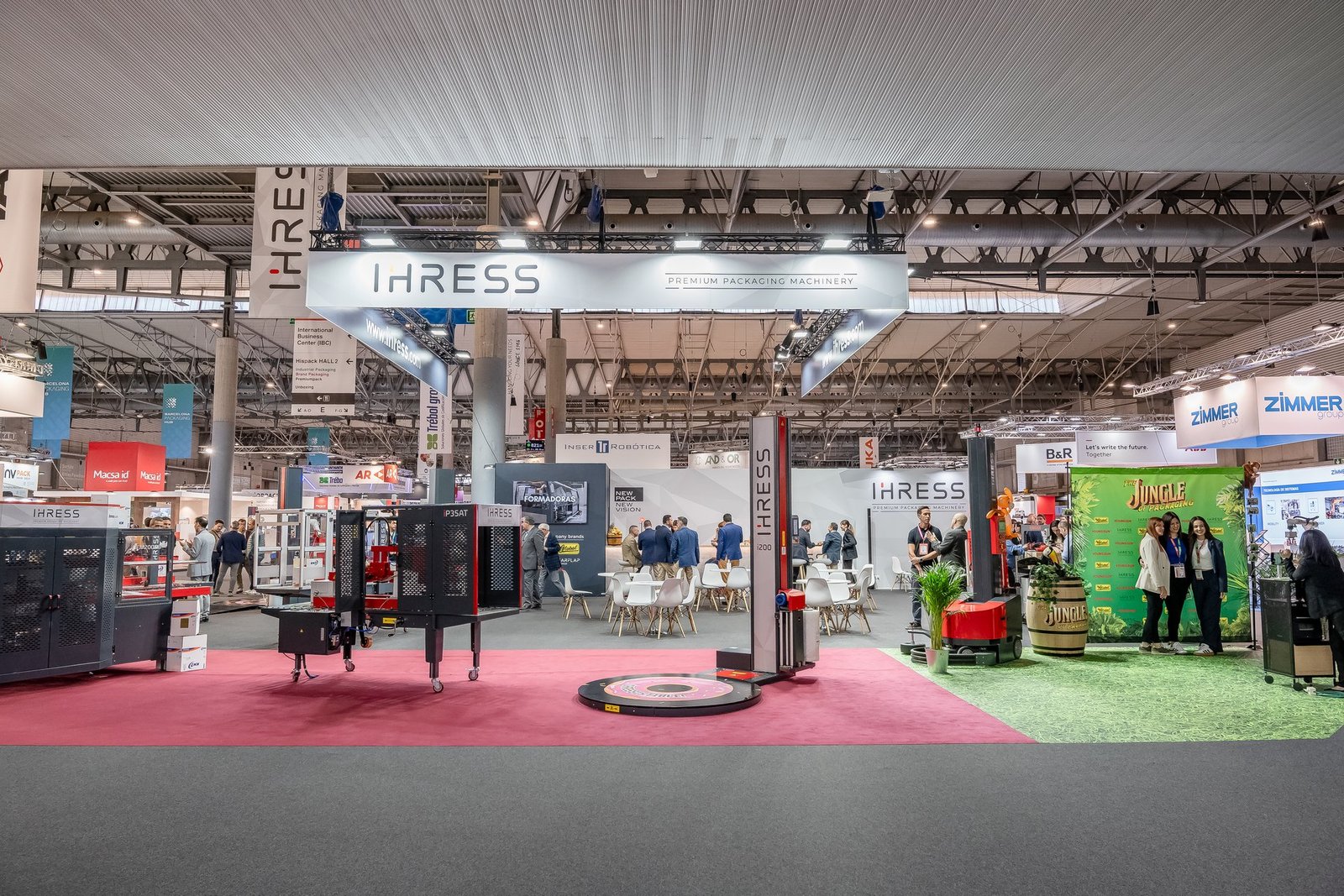 Stand de IHRESS en Hispack con maquinaria de embalaje y área de lounge. Visitantes interactúan con el personal y las máquinas de packaging.
