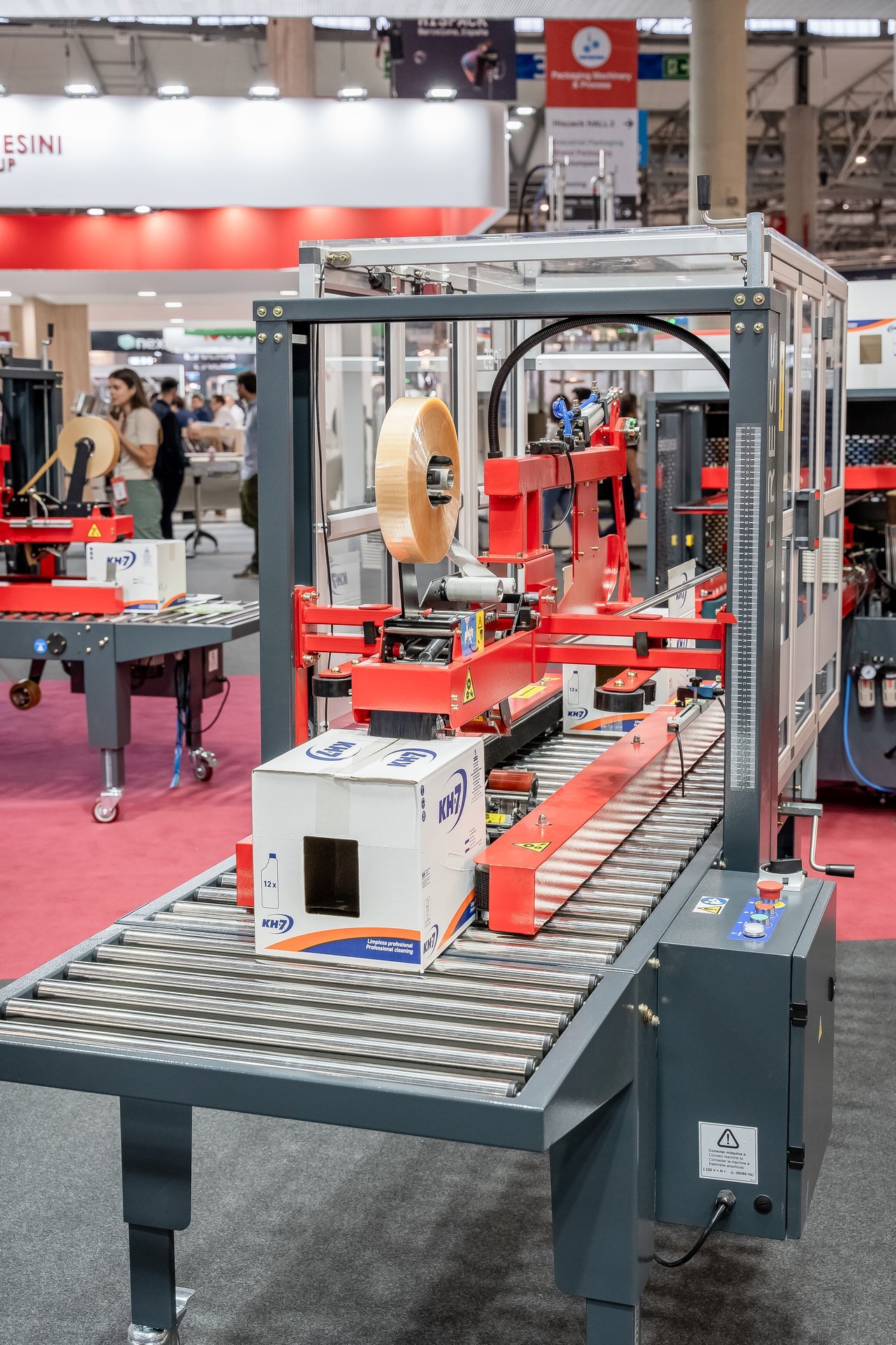 Máquina de embalaje automática sellando una caja blanca de KH7 en una feria industrial. Se ve un rollo de cinta adhesiva.