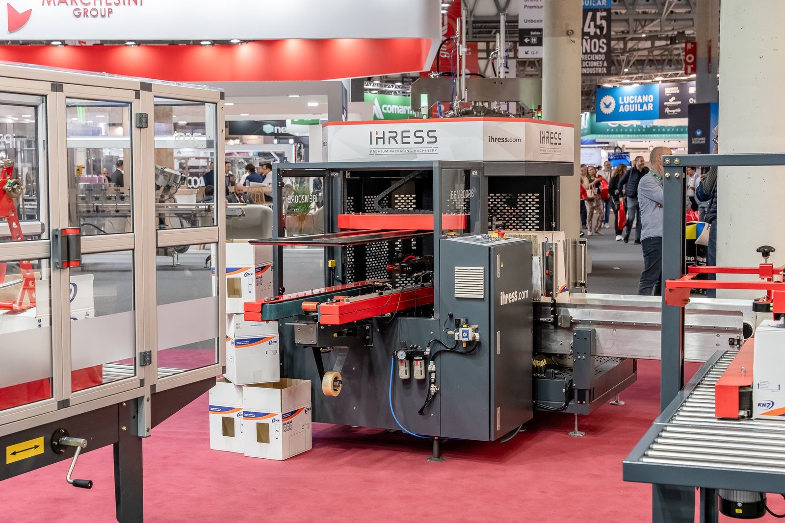 Máquina de embalaje automática IHRESS en la feria Hispack, con cajas de cartón apiladas y visitantes en el fondo.