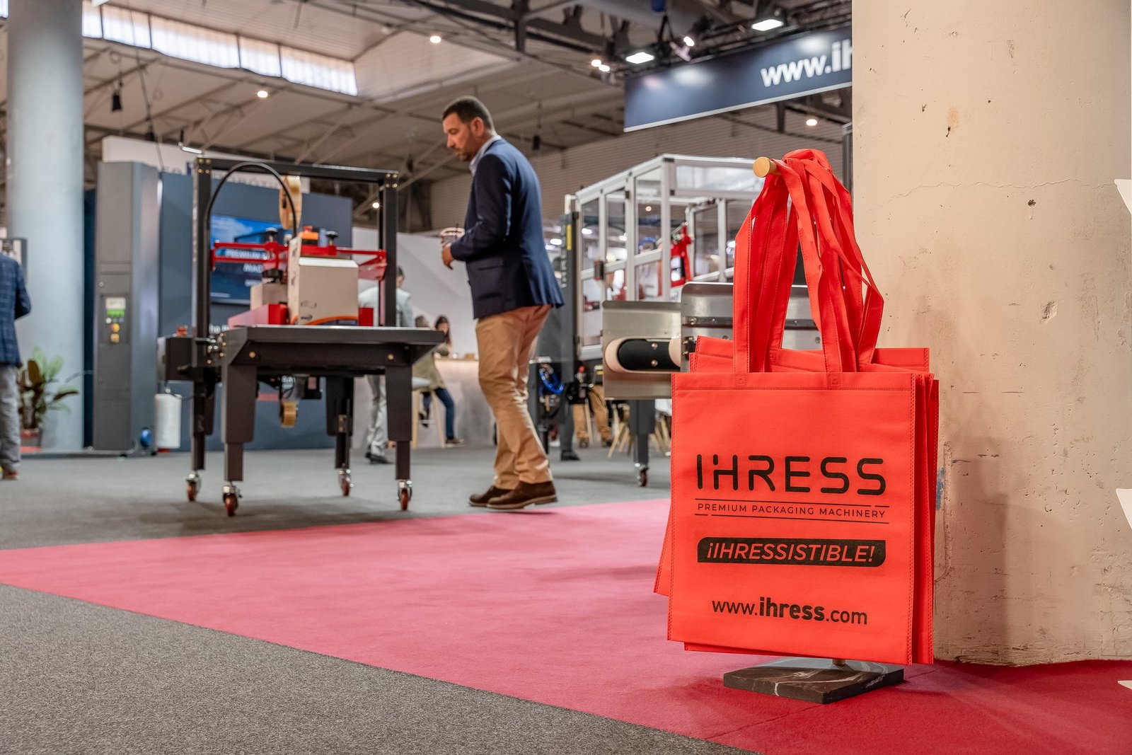 Bolsas naranjas de IHRESS en un stand de Hispack. Un hombre camina junto a maquinaria de embalaje en la feria.