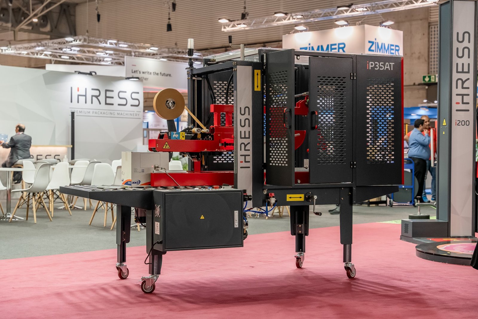 Máquina de embalaje IHRESS iP35AT negra y roja procesando una caja con cinta adhesiva en el stand de Hispack.