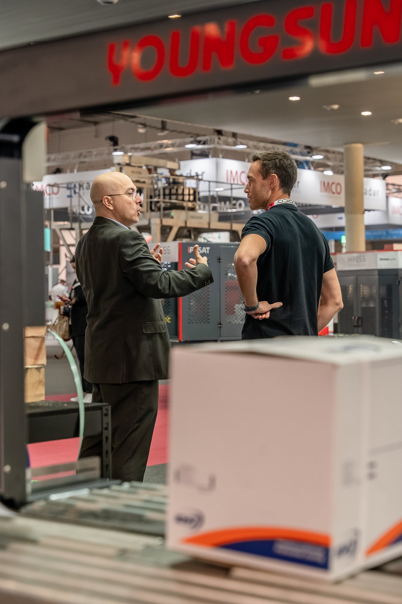 Dos hombres conversan en el stand de Hispack. Uno con traje gesticula, el otro de espaldas. Maquinaria industrial y cajas.