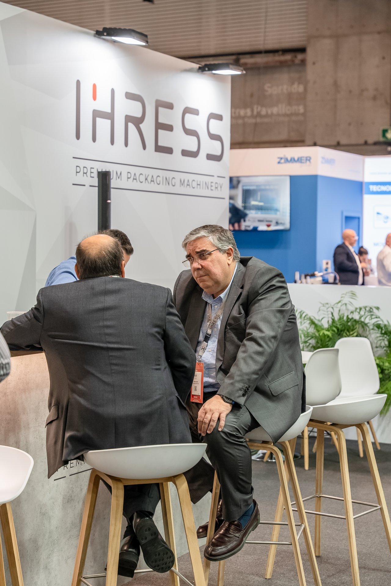 Dos hombres de negocios sentados en taburetes altos en el stand de IHRESS en la feria Hispack, conversando.