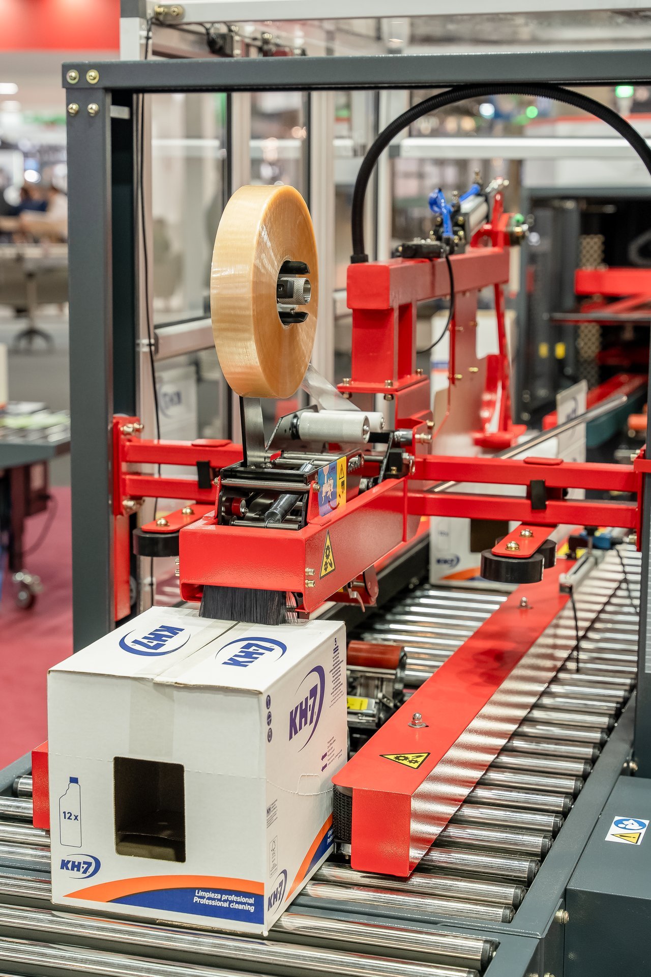 Máquina de embalaje sellando una caja de productos KH7 con cinta adhesiva en una feria industrial.
