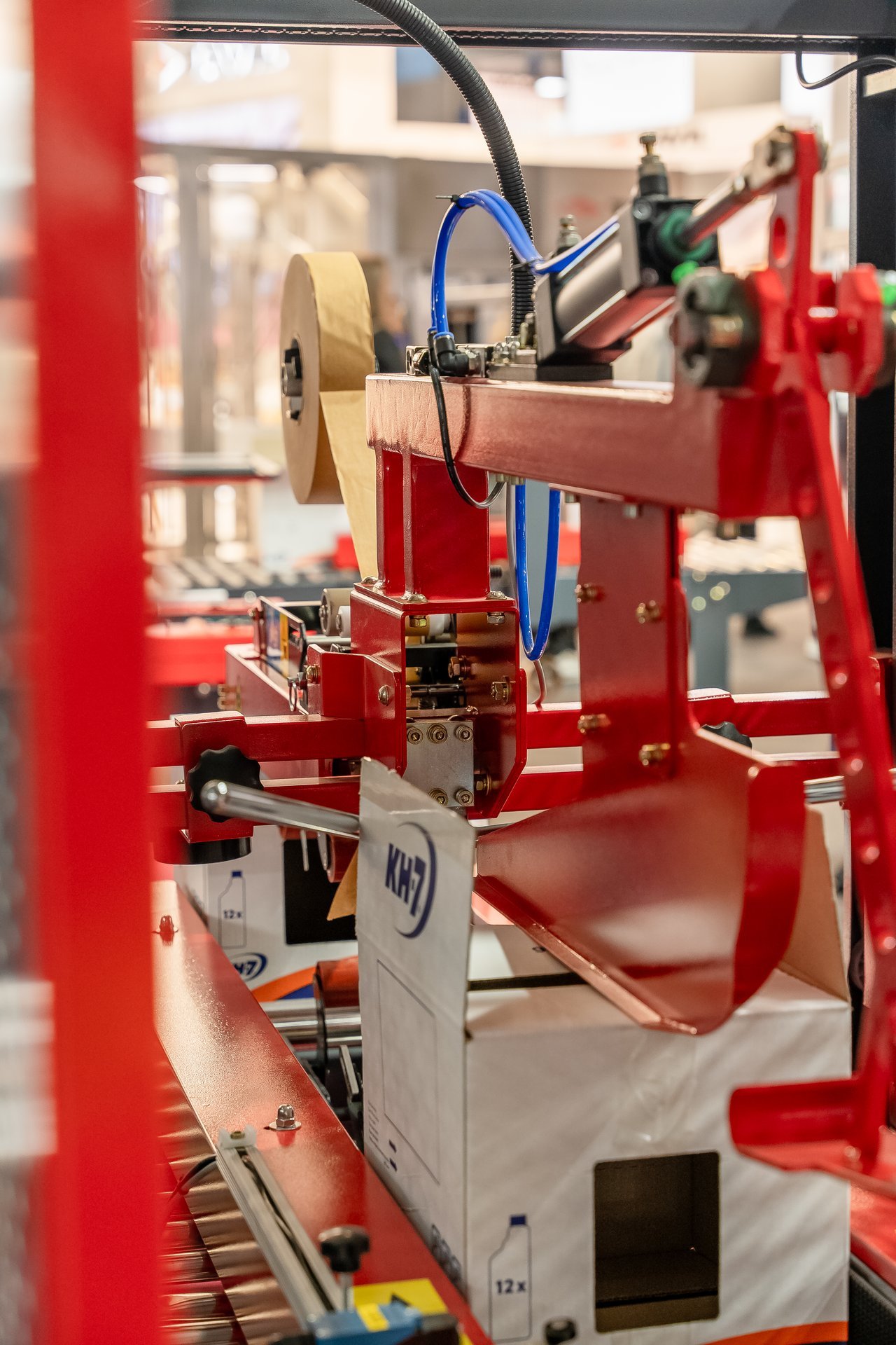 Máquina industrial roja con rollo de cinta adhesiva procesando una caja blanca con logo KH7. Evento Hispack.