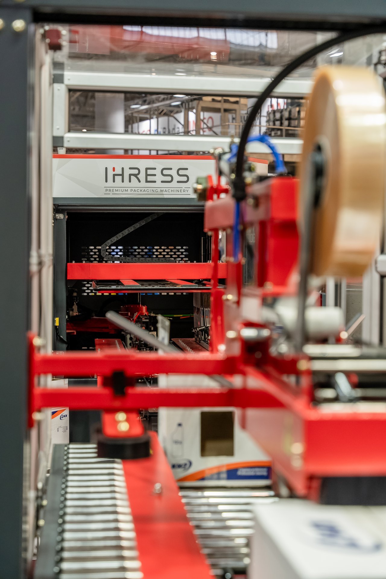 Máquina de embalaje IHRESS con logo 'Premium Packaging Machinery' en Hispack. Detalle de componentes rojos, cinta transportadora y rollo…