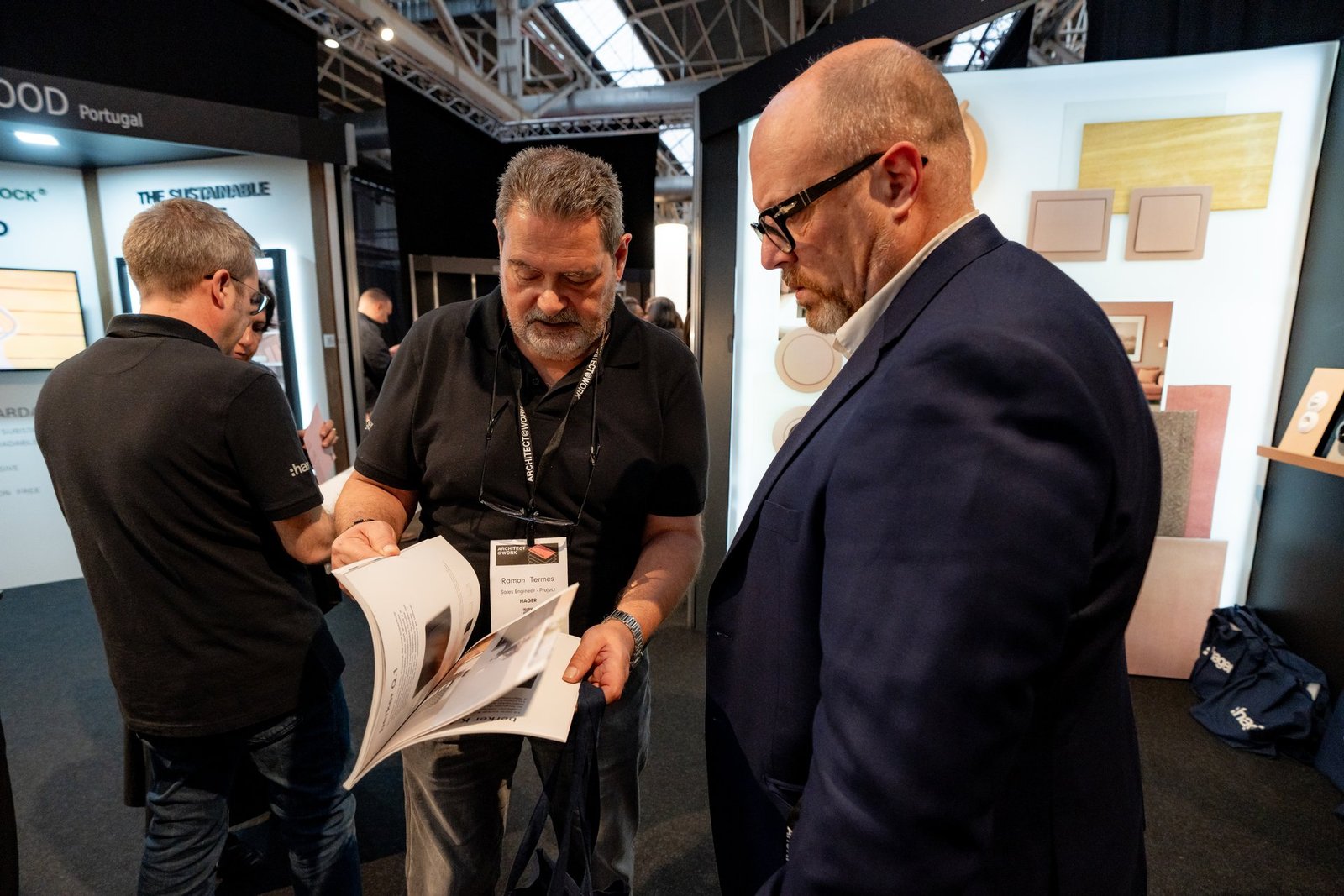 Dos hombres en Architect@Work, uno de Hager, revisan un catálogo en un stand de exposición. Fondo con paneles de materiales.