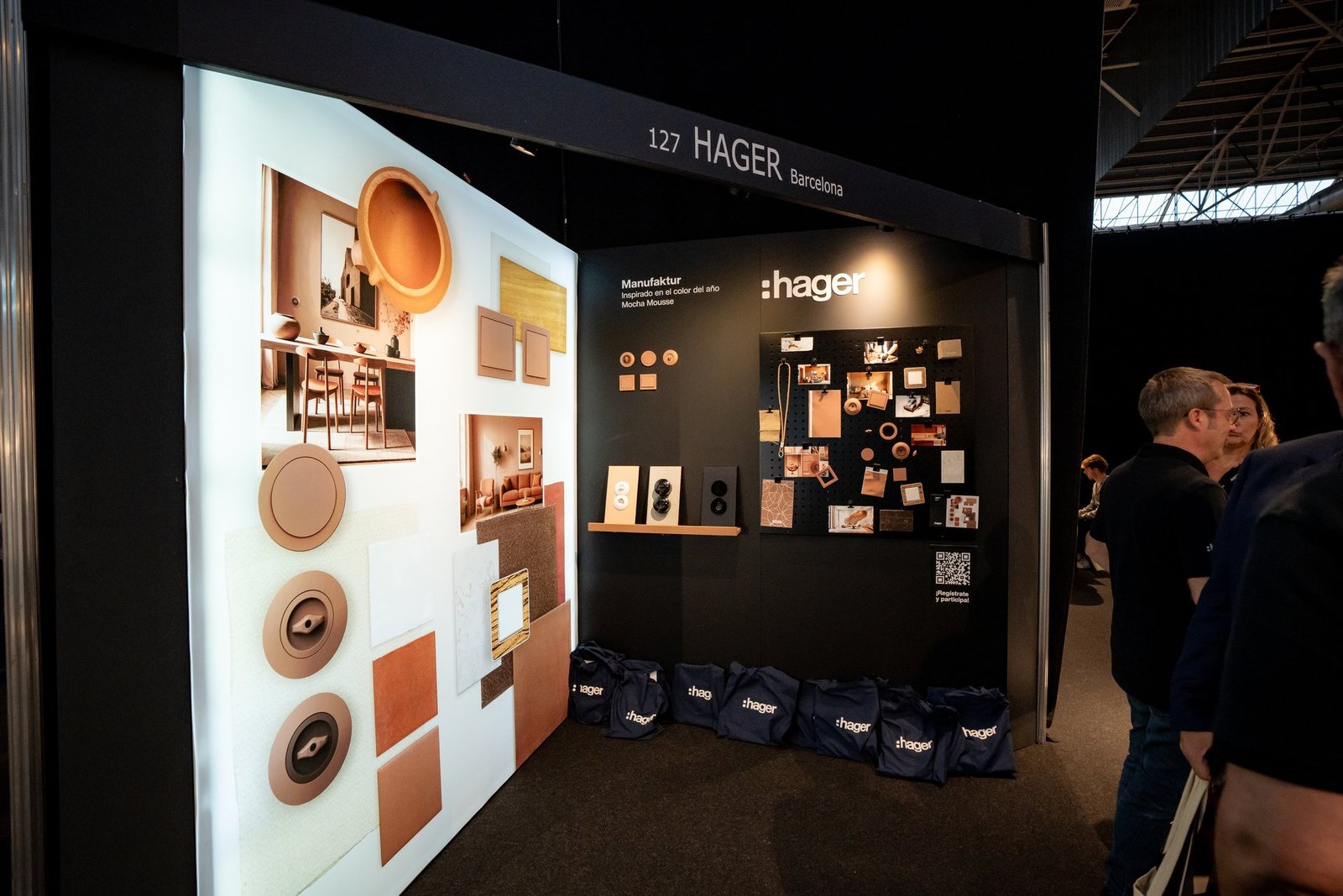 Stand de Hager en evento. Panel iluminado con productos y texturas en tonos tierra. Bolsas azules con logo Hager. Personas observando la…