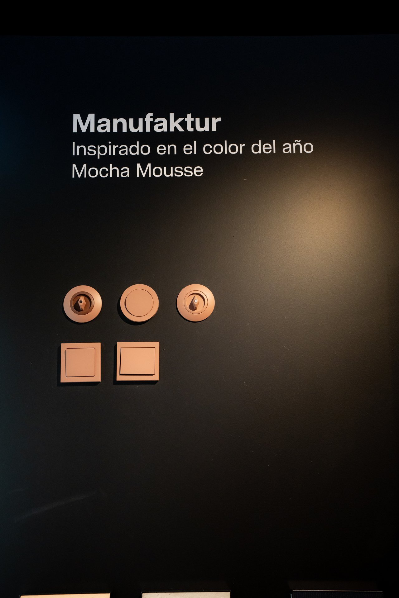 Texto 'Manufaktur, inspirado en el color Mocha Mousse'. Cinco interruptores y enchufes Hager color cobre en pared oscura.