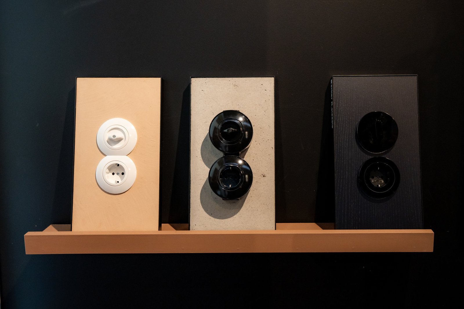 Exposición de interruptores y enchufes Hager: blancos en madera, negros en hormigón y negros en panel oscuro, sobre estante.
