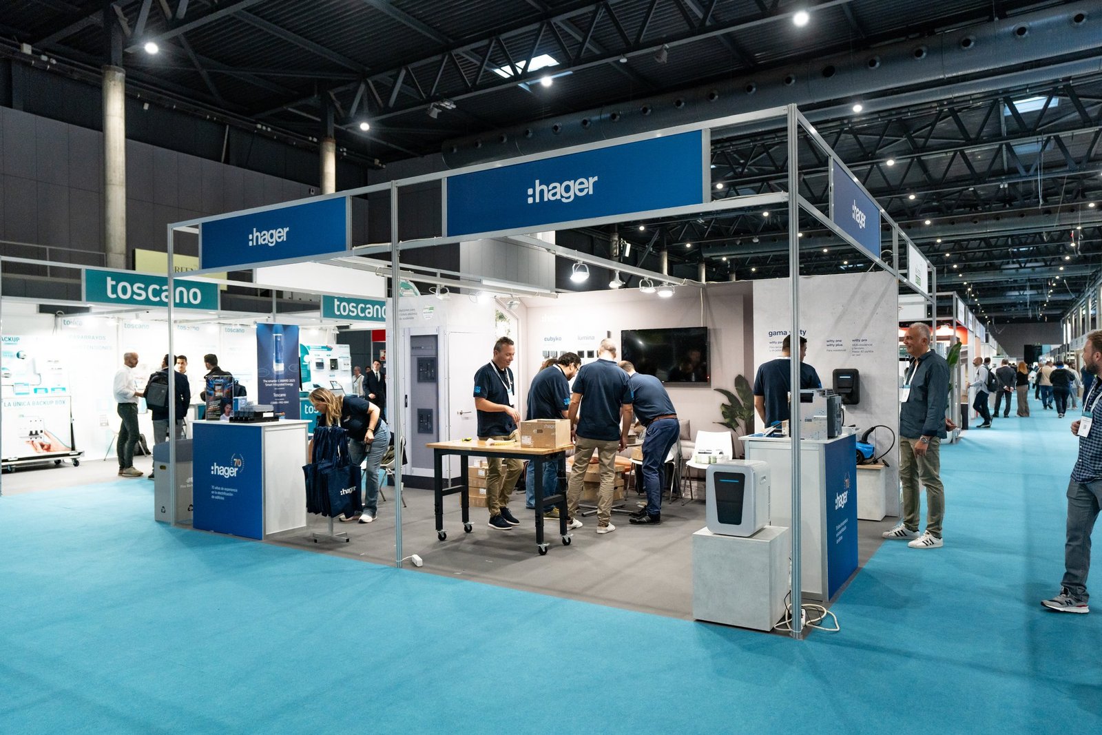 Stand de Hager en una feria, con personal y visitantes interactuando. Se ven productos eléctricos y el logo de Toscano al fondo.
