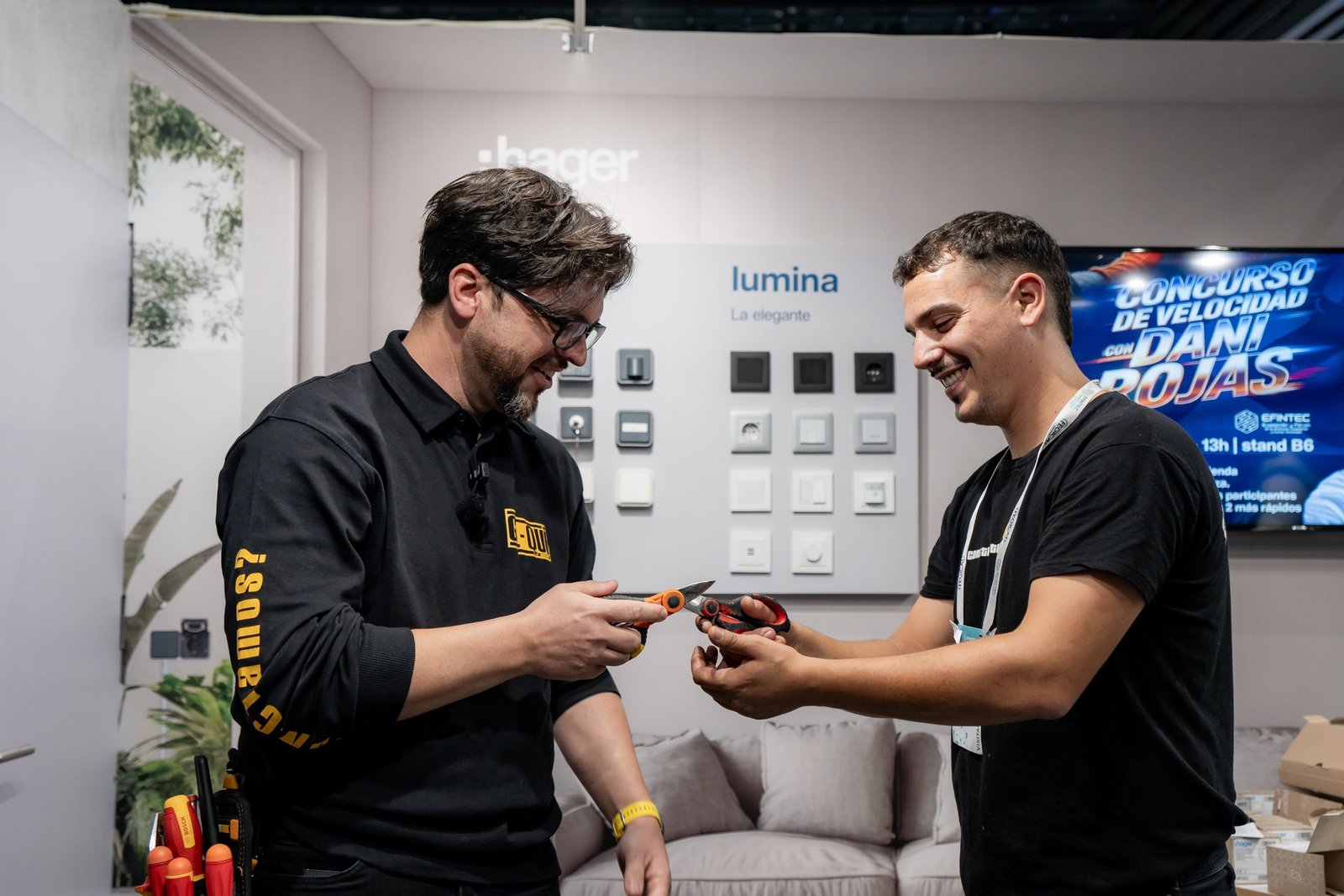 Dos hombres sonrientes intercambian tijeras en el stand de Hager, con una exhibición de interruptores y enchufes Lumina.
