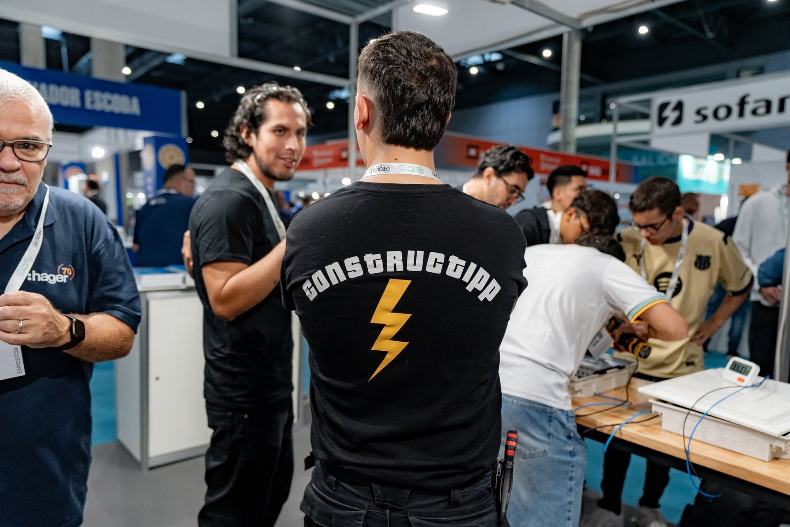 Asistentes en el evento Efintec de Hager. Un hombre con camiseta "CONSTRUGGIPP" de espaldas, y otros interactuando en un stand.
