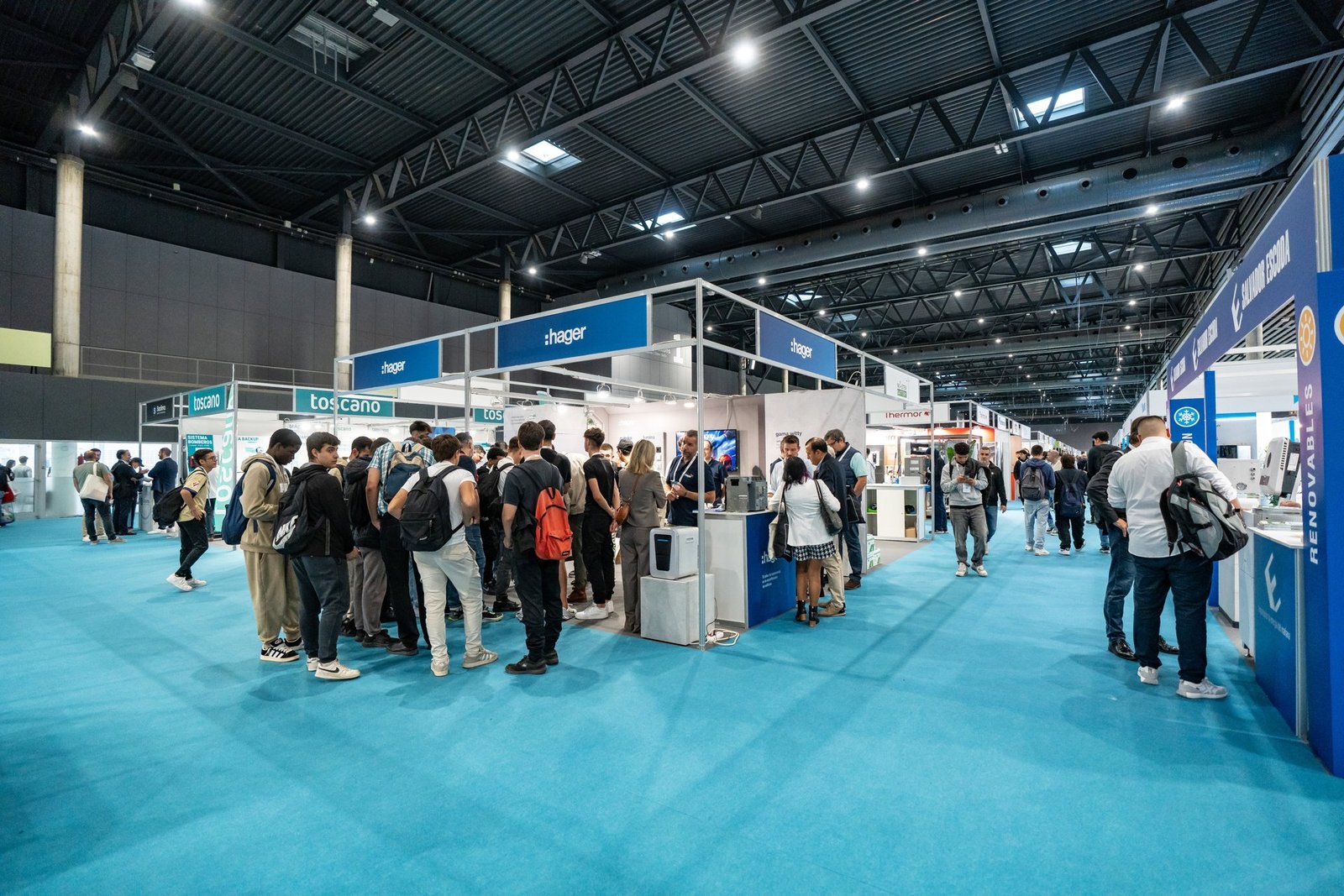 Pabellón de exposición concurrido en Efintec. Gente interactuando en stands de Hager y Toscano. Moqueta azul, techo alto.