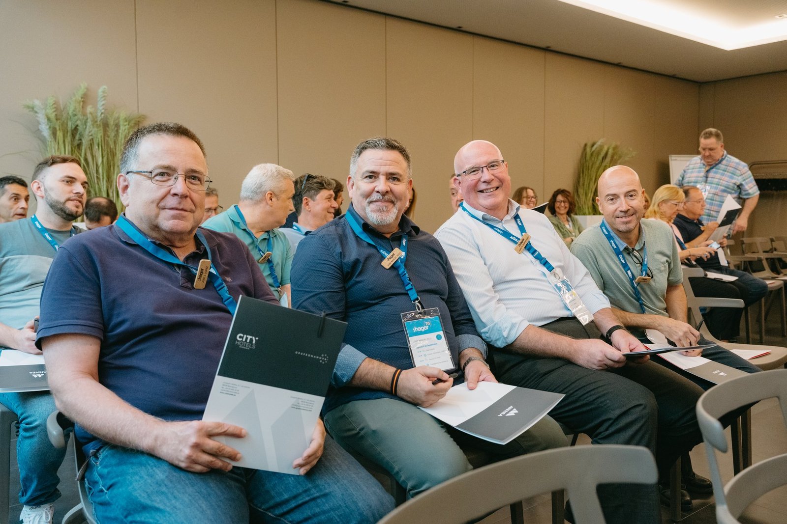 Grupo de asistentes sonrientes en un evento teambuilding de Hager. Llevan lanyards y sostienen documentos en una sala de conferencias.