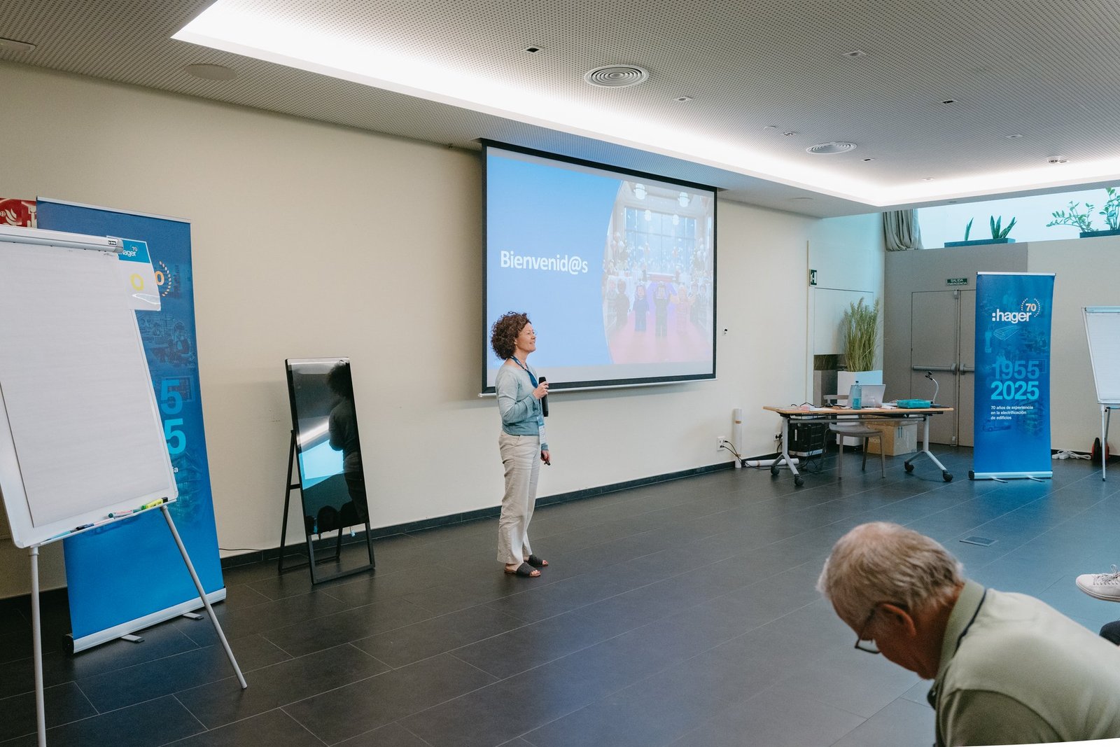 Mujer con micrófono presentando en un evento de Hager. La pantalla proyecta "Bienvenid@s" y una imagen de grupo. Hay banners de Hager.
