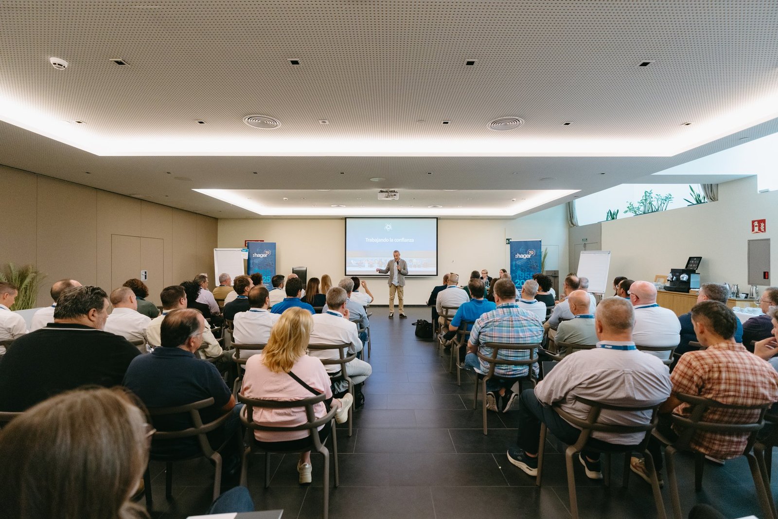 Un orador presenta a una audiencia sentada en un evento de teambuilding de Hager. La pantalla muestra 'Trabajando la confianza'.