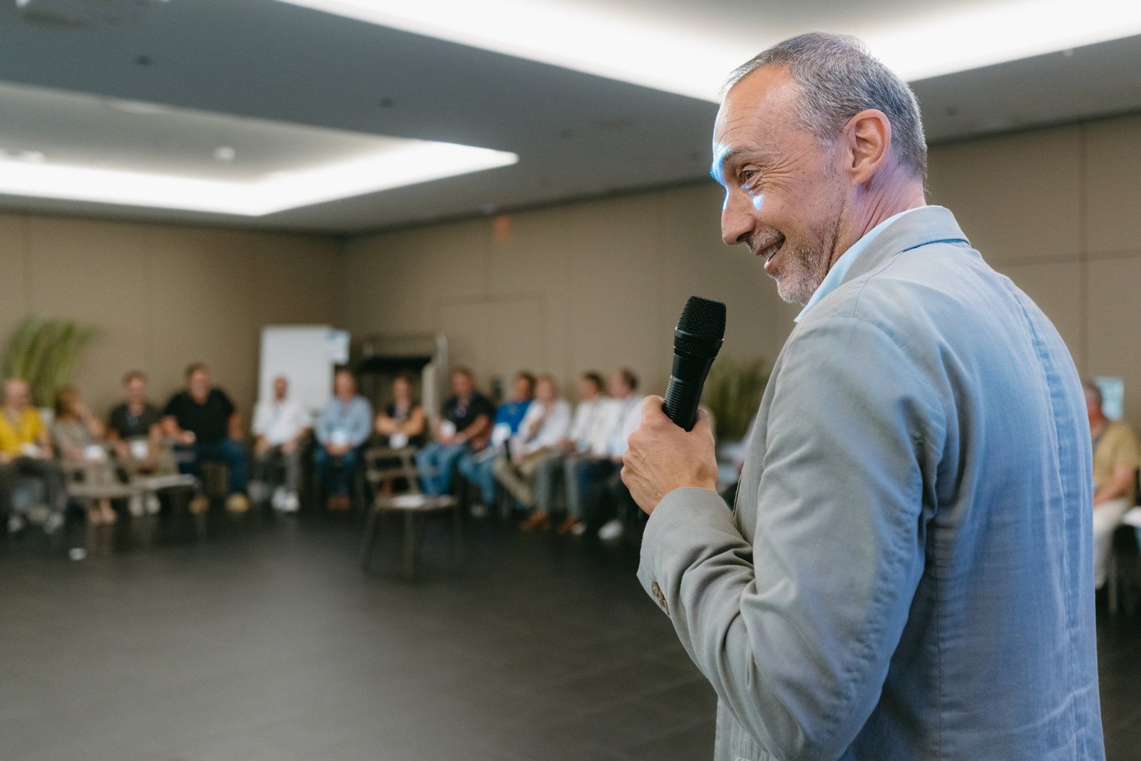 Hombre sonriente con micrófono, dirigiéndose a un grupo de personas sentadas en semicírculo en un evento.