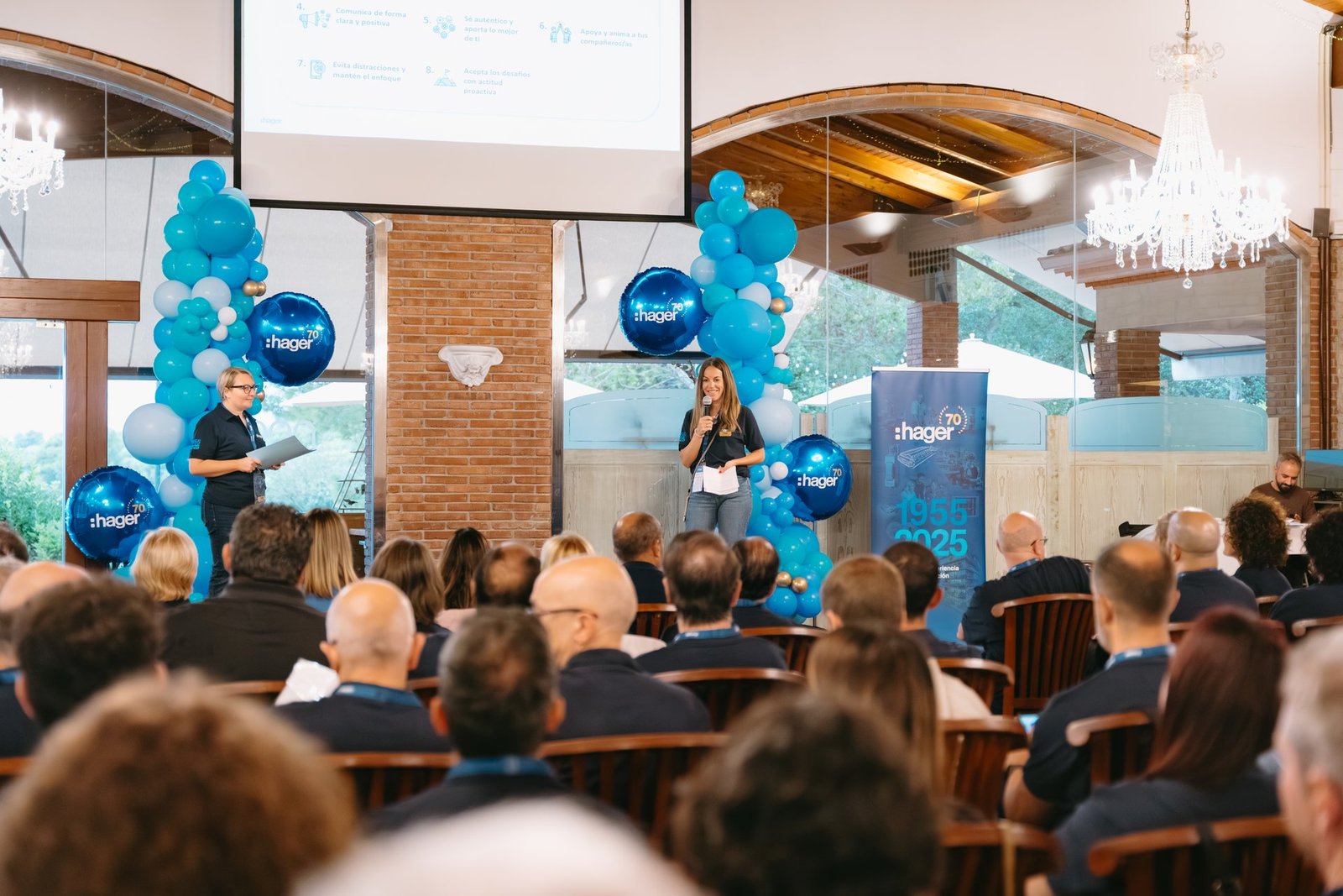 Dos mujeres presentan en un evento de teambuilding de Hager. Una habla por micrófono, la otra sostiene papeles. Audiencia sentada, globos…