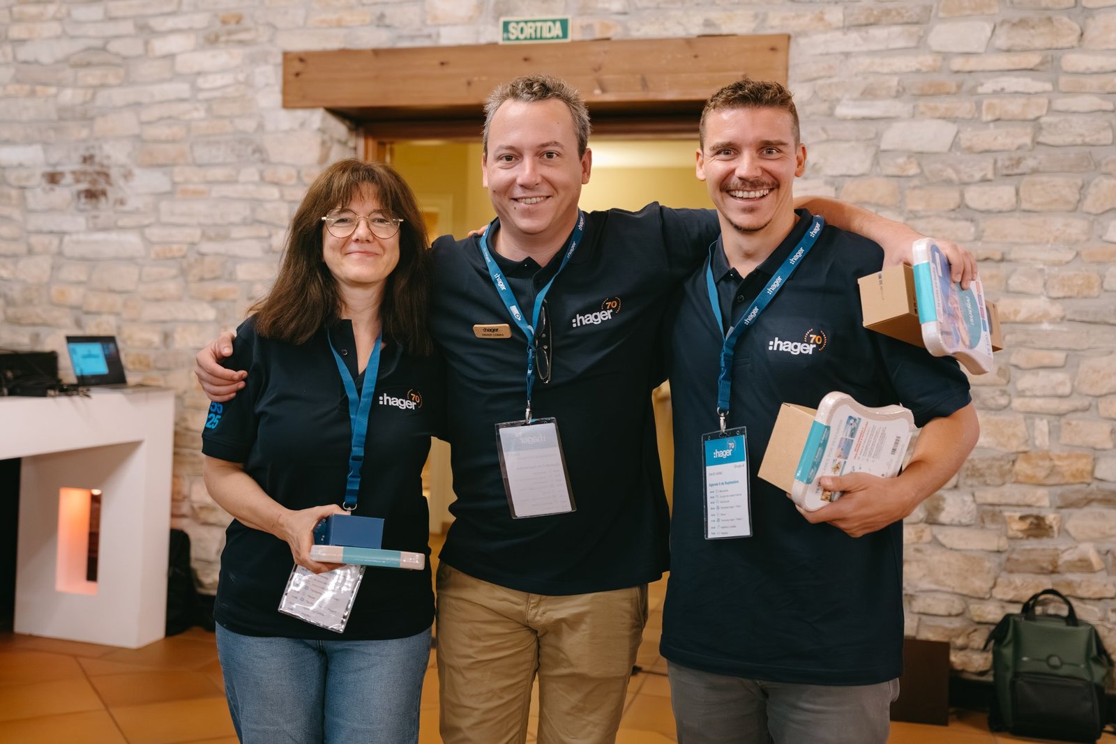 Tres empleados de Hager sonríen, con polos azules y regalos, en un evento de teambuilding. Fondo de pared de piedra.