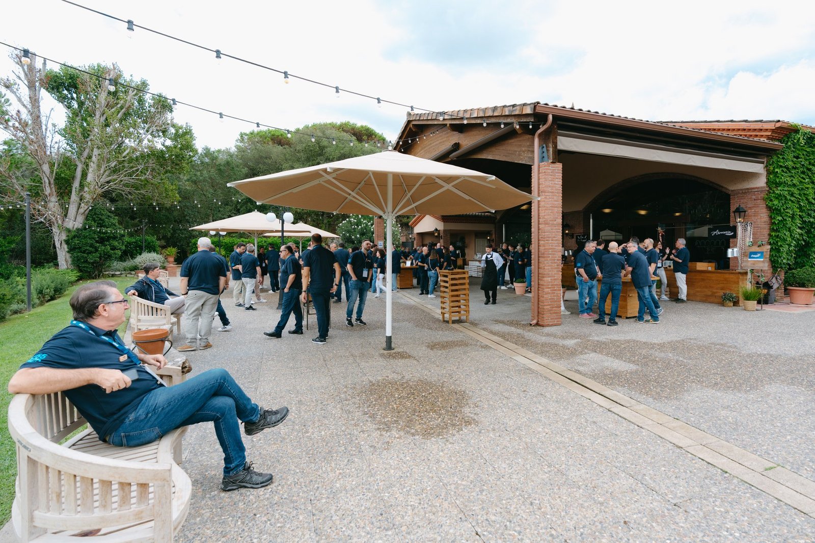 Grupo de personas en un evento de teambuilding al aire libre. Hombres con polos azules conversan en un patio con sombrillas y un edificio.