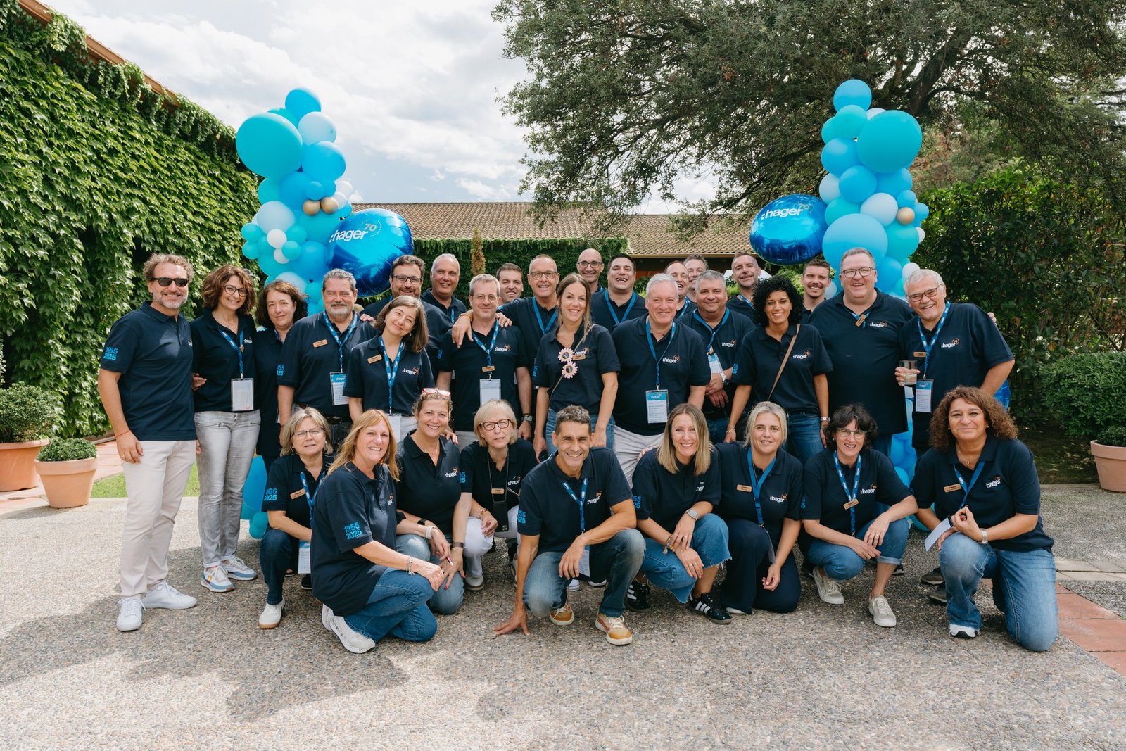Grupo grande de empleados de Hager sonriendo, posando al aire libre con polos azules. Globos azules decoran el fondo.