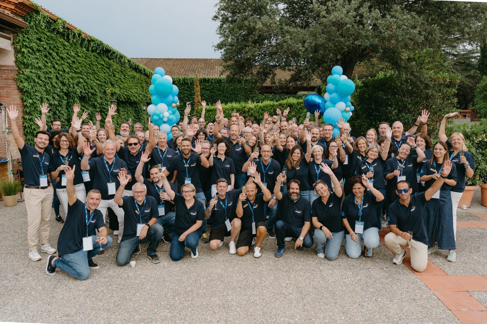 Gran grupo de empleados de Hager sonriendo y saludando en un evento de teambuilding. Visten polos azules y hay globos decorativos.