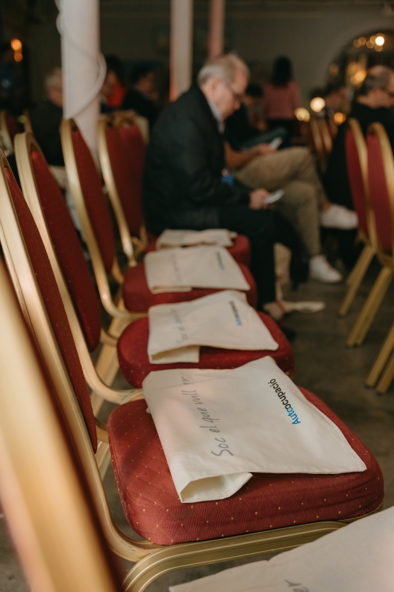 Filas de sillas rojas con bolsas blancas de Autoocupació, algunas con el lema "Soc el que vull ser". Asistentes al evento.