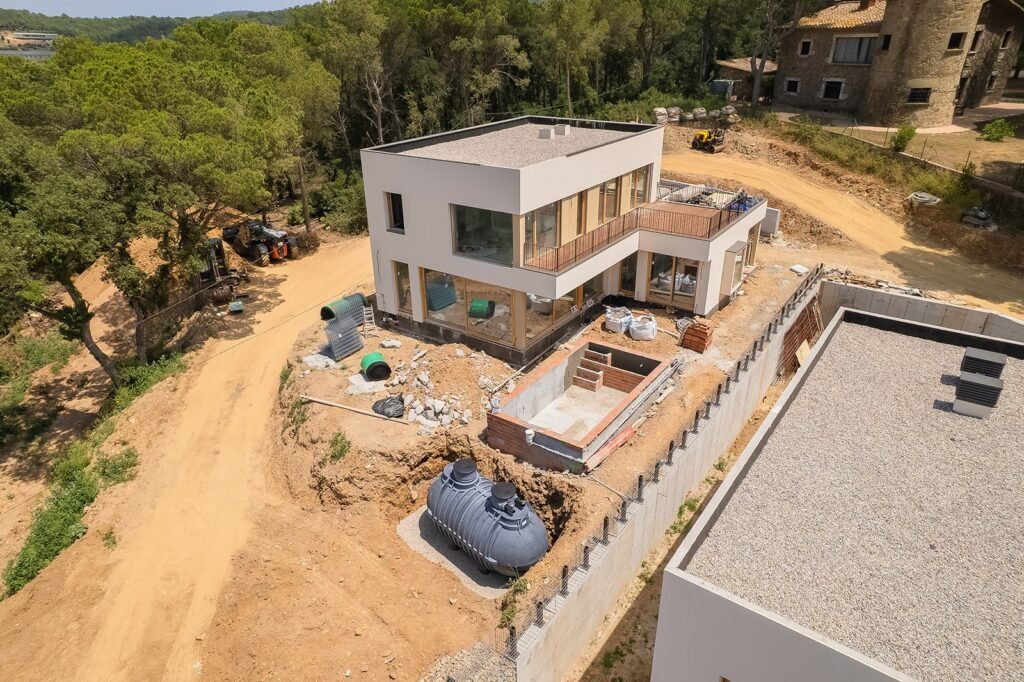 Vista aérea de obra: casa moderna en construcción con piscina, tanque subterráneo y excavaciones. Maquinaria en camino de tierra y bosque.