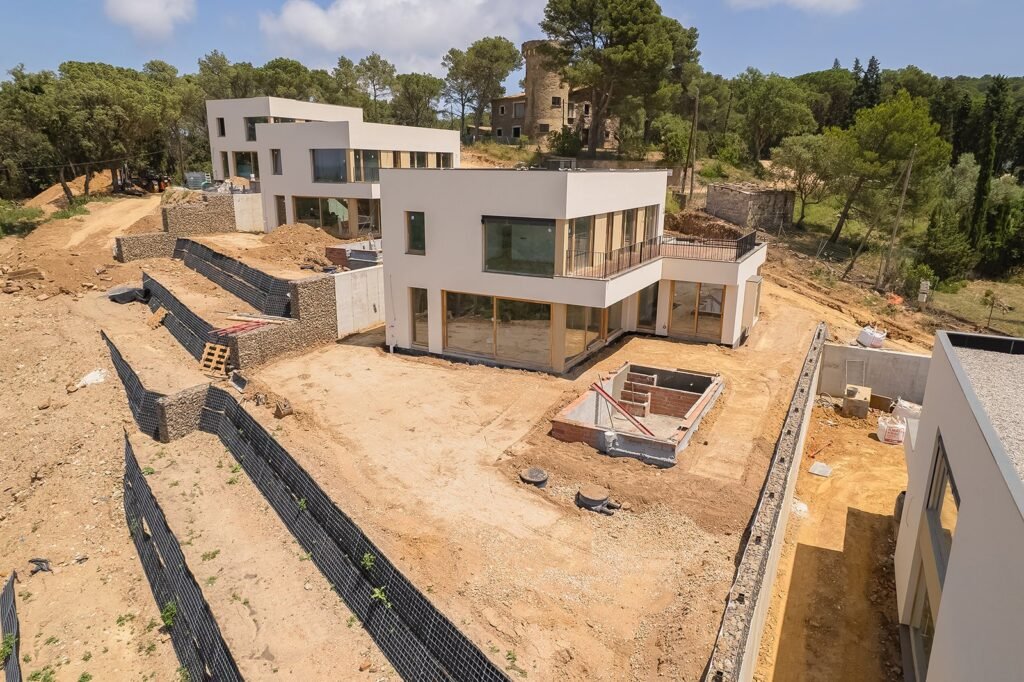 Vista aérea de casas modernas en construcción con fachadas blancas y grandes ventanales. Terreno inclinado con muros de gaviones y bosque…