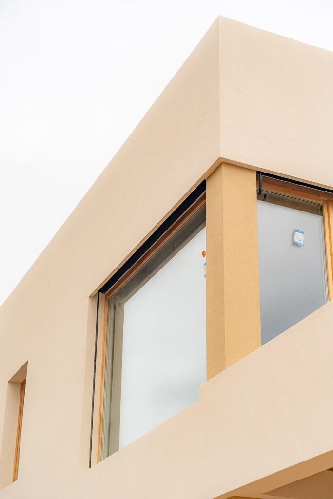 Fachada de edificio moderno en construcción con acabado beige claro. Grandes ventanales y diseño minimalista de ángulos rectos.