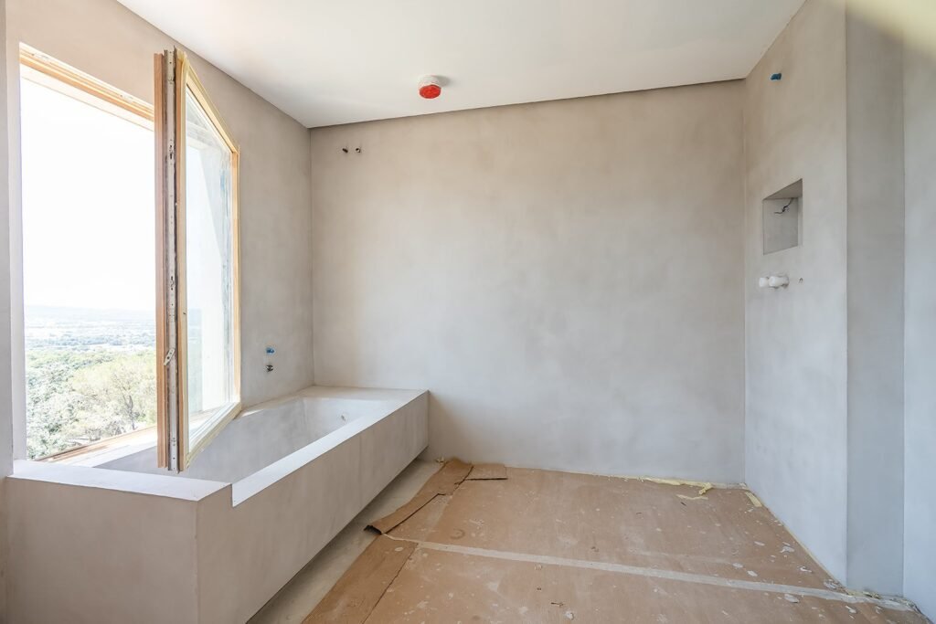 Baño en construcción con bañera de obra, ventana abierta mostrando vistas al exterior y paredes enlucidas.