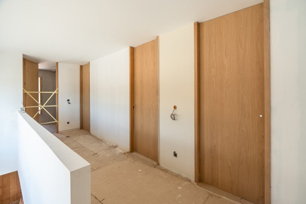 Pasillo de obra con puertas de madera, paredes claras y suelo protegido. Barandilla blanca a la izquierda. Cables eléctricos expuestos.