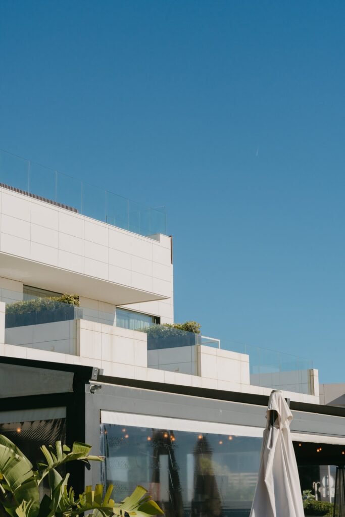 Edificio moderno blanco con balcones de cristal y plantas, y parte de una terraza con sombrilla plegada bajo cielo azul.
